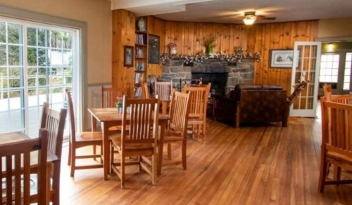 Cozy interior of a rustic dining area with wooden tables, chairs, and a stone fireplace. Natural light from windows.