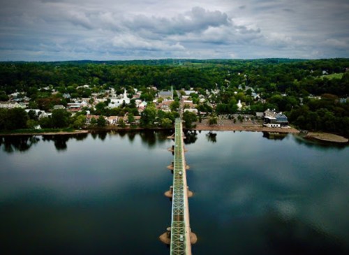 Aerial view of a bridge spanning a calm river, with a small town and lush greenery in the background under cloudy skies.