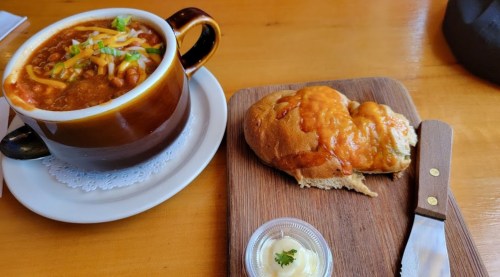 A bowl of chili topped with cheese and green onions, alongside a cheesy bread roll and a small dish of butter.