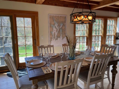 A cozy dining room with a wooden table set for dinner, featuring a map on the wall and large windows overlooking greenery.