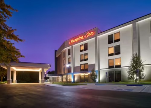 A Hampton Inn hotel exterior at dusk, featuring illuminated signage and modern architecture.