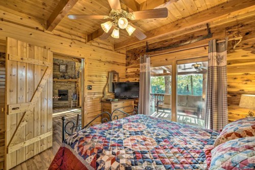 Cozy wooden bedroom with a quilted bedspread, ceiling fan, and sliding door leading to a deck with forest views.