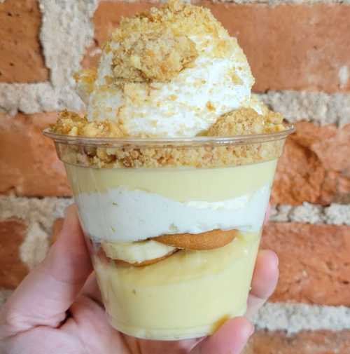 A hand holding a clear cup filled with layered banana pudding, whipped cream, and crushed cookies against a brick wall.