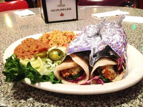 A plate with two foil-wrapped tacos, rice, refried beans, lettuce, jalapeños, and lime on a speckled table.