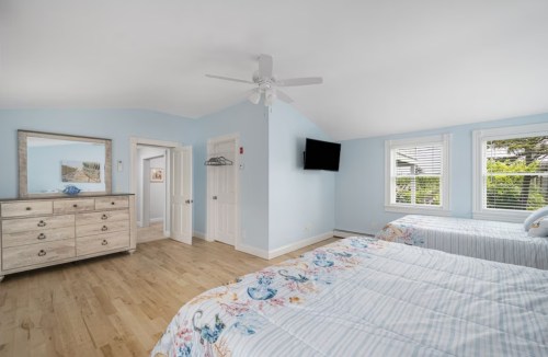 Bright bedroom with two beds, light blue walls, a dresser, and a TV, featuring large windows and wooden flooring.