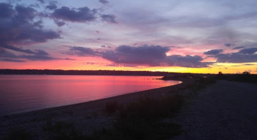 A serene sunset over a calm lake, with vibrant pink and purple hues reflecting on the water and a rocky shoreline.