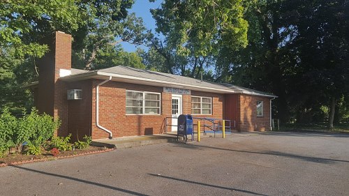 A brick building with a blue mailbox outside, surrounded by trees and greenery, in a sunny setting.