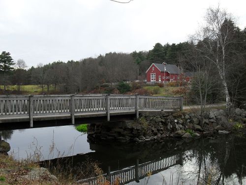 A wooden bridge spans a calm pond, with a red house and trees in the background under a cloudy sky.