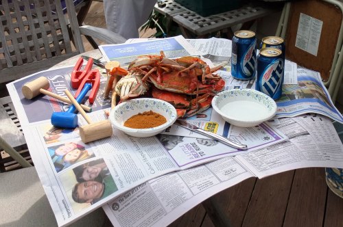 A table set with crabs, seasoning, tools, and drinks, all laid out on newspapers for a seafood feast.