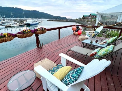 A serene waterfront view from a deck with chairs, colorful cushions, and flower baskets overlooking a marina.