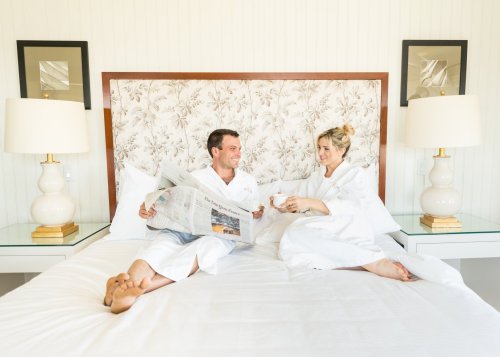 A couple in white robes sits on a bed, smiling and reading a newspaper together, with lamps and framed art in the background.