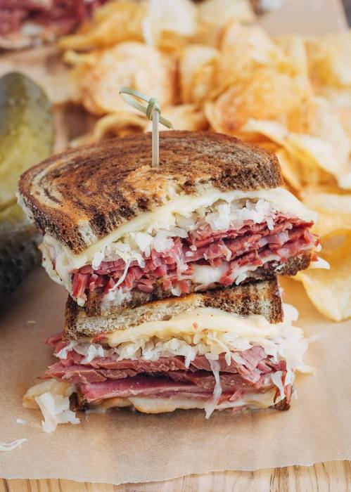 A stacked sandwich with layers of corned beef, sauerkraut, and cheese, served with chips and pickles on the side.