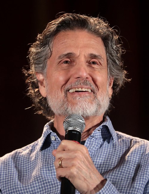 A smiling man with curly gray hair and a beard holds a microphone, wearing a blue checkered shirt.