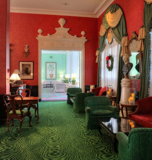 Elegant room with red walls, green carpet, and vintage furniture, featuring a doorway to a bright, decorated space.