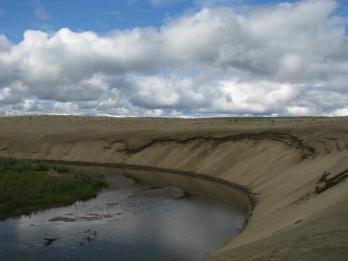 A winding river flows through a sandy landscape under a cloudy sky, with green grass peeking through the dunes.