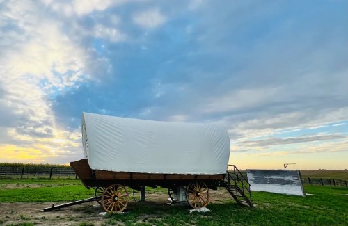 A covered wagon on a grassy field under a cloudy sky at sunset, with a wooden ramp beside it.