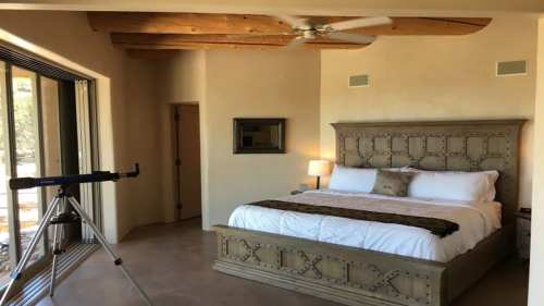 A cozy bedroom with a large bed, wooden beams, and a telescope by the window, featuring warm, neutral tones.