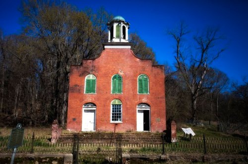 Historic brick building with green shutters and a cupola, surrounded by trees under a clear blue sky.