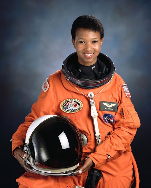 A smiling astronaut in an orange space suit holds a helmet, set against a blue background.