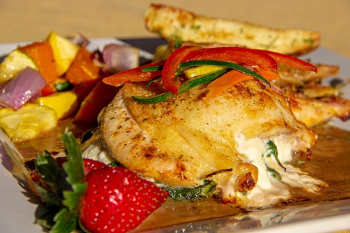A plated dish featuring grilled chicken with vegetables, a side of fruit salad, and a strawberry garnish.