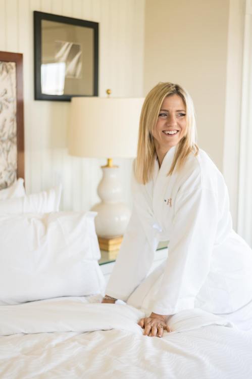 A woman in a white robe smiles while adjusting bedding in a bright, cozy room.
