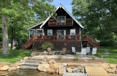 A charming brown cabin by a lake, featuring a deck, patio furniture, and surrounded by lush greenery.