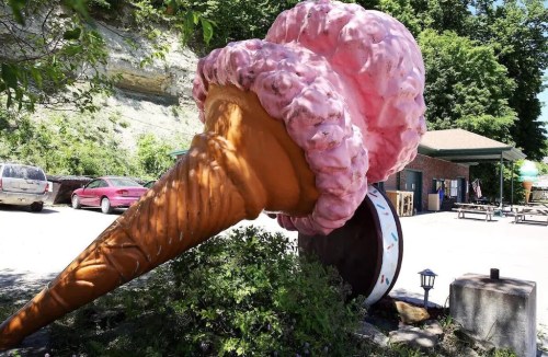 A large, whimsical ice cream cone sculpture with pink ice cream and a brown cone, set in a grassy area.