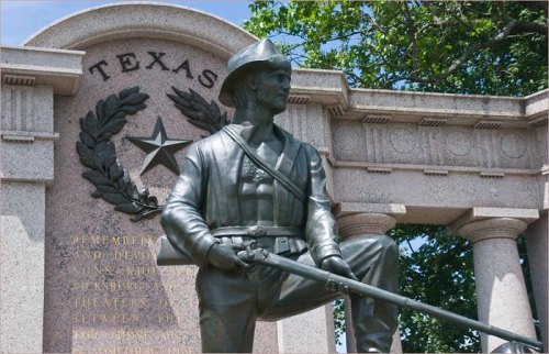 Statue of a soldier in front of a monument with a Texas star and inscriptions, surrounded by trees.