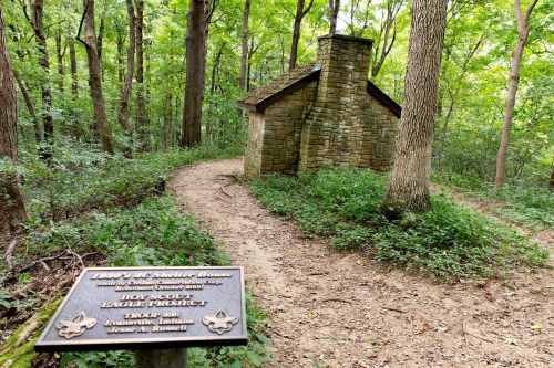 A stone shelter house in a wooded area, with a plaque in the foreground detailing its history and significance.