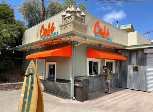 A café named "Sand Castle" with orange awnings, a castle-like structure on top, and a man ordering at the window.