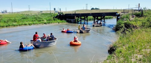 People enjoying a sunny day on a river, floating in boats and inner tubes near a wooden bridge.