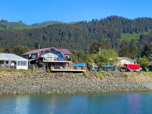 A scenic view of a house by the water, surrounded by trees and mountains under a clear blue sky.