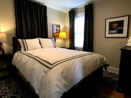 Cozy bedroom with a queen bed, dark curtains, a lamp, and framed artwork on the walls.