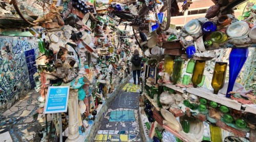 A colorful, eclectic pathway lined with bottles and art, leading through a whimsical, decorated space.
