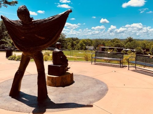 A bronze statue of a man in a cape stands beside a seated figure, overlooking a park with blue skies and greenery.