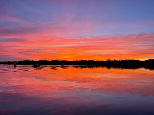A serene lake at sunset, reflecting vibrant pink and orange hues in the sky and water, with boats in the distance.
