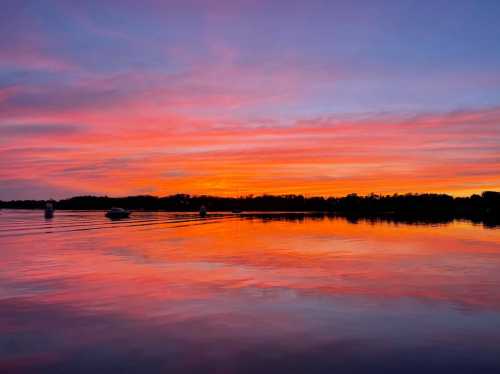 A serene lake at sunset, reflecting vibrant pink and orange hues in the sky and water, with boats in the distance.