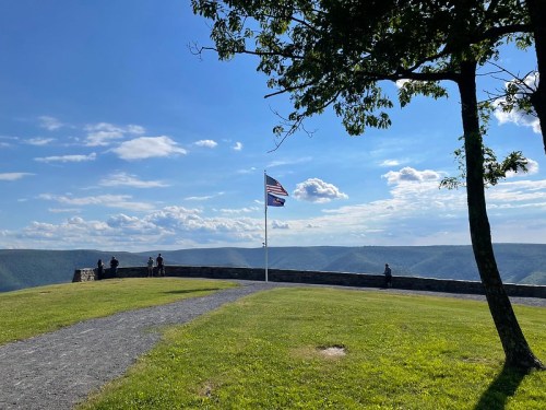 A scenic view from a stone overlook with a flag, surrounded by green grass and distant mountains under a blue sky.
