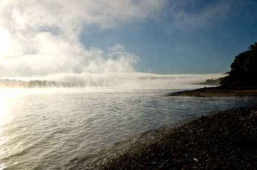 A serene river scene with mist rising over the water, framed by trees and a clear blue sky.