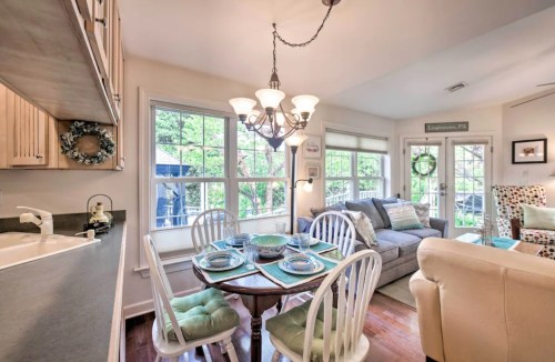 Cozy dining area with a round table, chairs, and a chandelier, adjacent to a living space with a sofa and large windows.
