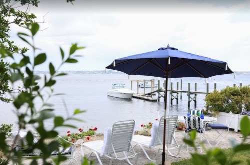 A serene waterfront scene with lounge chairs, a blue umbrella, and a boat docked nearby under a cloudy sky.