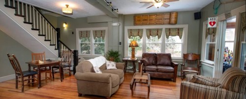 Cozy living room with a sofa, armchairs, dining table, and large windows, featuring wooden floors and a staircase.