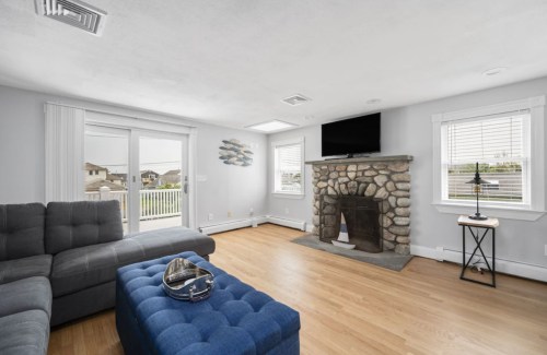 Bright living room with a stone fireplace, gray sectional sofa, blue ottoman, and large windows overlooking a deck.