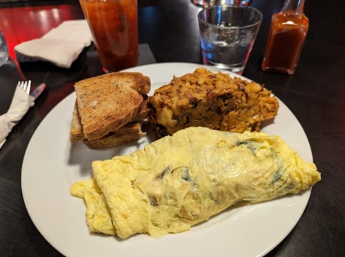 A plate with a fluffy omelette, hash browns, toast, and a glass of water, set on a dark table.