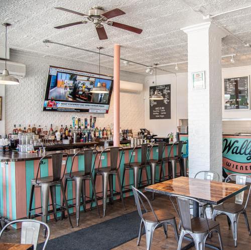 A vibrant bar area with a TV, colorful decor, and metal stools, featuring a wooden table and a welcoming atmosphere.