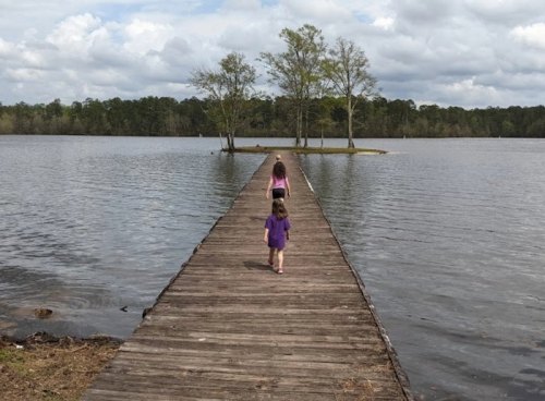 Two children walk along a wooden pier extending into a calm lake, surrounded by trees and cloudy skies.