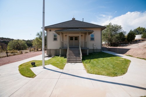 A light-colored building with a porch, surrounded by grass and a concrete pathway, under a clear blue sky.