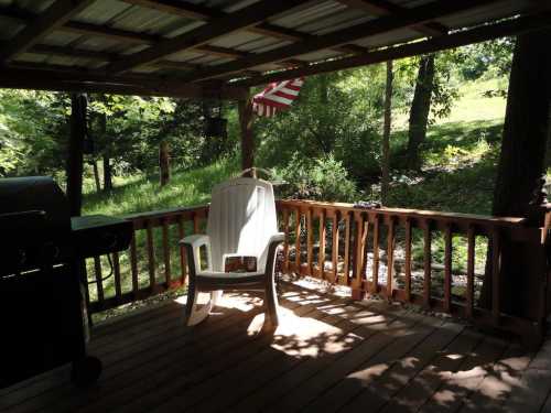 A shaded wooden deck with a white chair, a grill, and a view of greenery and trees in the background.