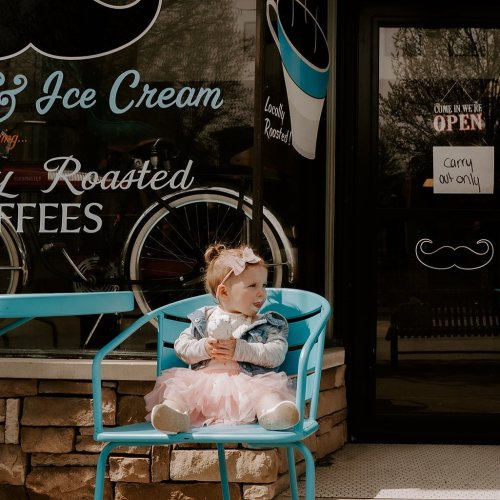 A young child in a pink tutu sits on a blue chair outside an ice cream shop, smiling and holding her hands together.