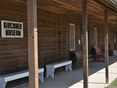 Exterior of the Kirchner Museum, featuring a wooden facade, benches, and a sign, with sunlight casting shadows.
